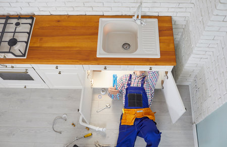 Plumber working in a modern kitchen. Plumber in an overall uniform lying down on the floor under the sink drain, repairing pipes or changing a rubber gasket. Top view, overhead, from above, high angleの写真素材