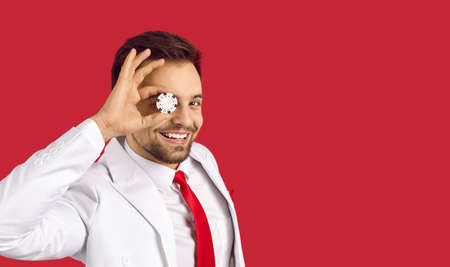 Happy lucky confident handsome young man in elegant white suit and tie covering one eye with poker chip in his hand standing on red blank text copyspace studio background. Casino and gambling conceptの写真素材