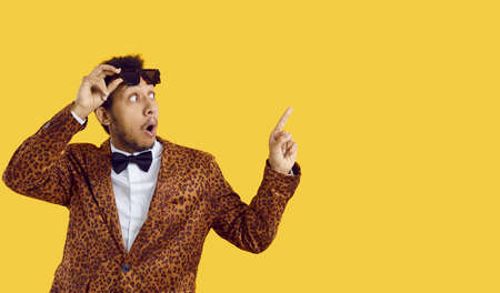 Surprised Afro American man in funky outfit and bowtie takes off his glasses and points index finger...