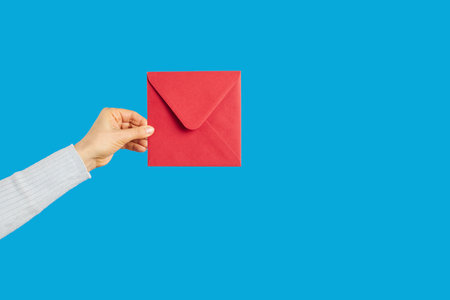 Cropped shot of female hand holding square red paper envelope against blue studio background. Courier, carrier, messenger, mailwoman delivering important letter, message or notice. Mail conceptの写真素材