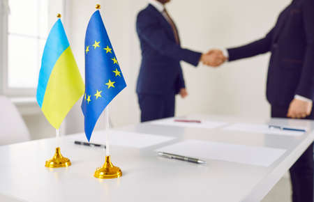 Flags of Ukraine and EU against background of representatives of countries shaking hands. Diplomats forge partnerships between countries. Concept of Ukraines accession to European Union. Close up.の写真素材