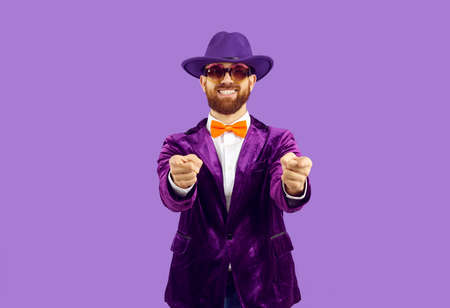 Portrait of excited male entertainer in velvet suit isolated on purple studio background point at screen. Smiling disco man or performer show at camera make choice. Choose you hand gesture.の写真素材
