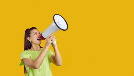 Happy confident beautiful young woman standing on bright yellow colour text copy space studio background, shouting through megaphone and making loud announcement about upcoming holiday sale eventの写真素材