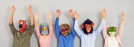 Portrait of unidentified young people in various rubber masks in form of animal heads. Young men and women in masks of horse, pigeon, dinosaur, monkey and frog stand in row with arms raised. Banner.の写真素材