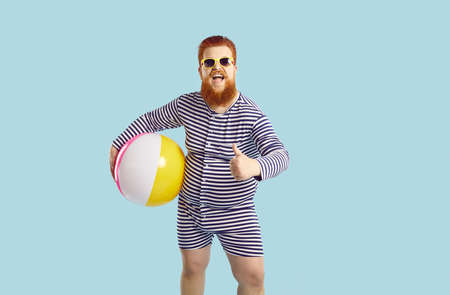 Funny cheerful chubby guy holds beach inflatable ball and shows thumb up on pastel turquoise background. Humorous redhead bearded young man in striped leotard recommends. Concept of summer vacation.の写真素材