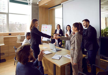 Two smiling women shaking hands while their teammates are applauding. Two teams of happy business people meeting in the office, making a deal, celebrating the start of collaboration and having funの写真素材