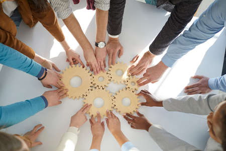 Top view close up of multiracial diverse businesspeople connect cogwheels work together for shared result. Employees join wheel gears involved in teambuilding at workplace. Teamwork concept.の写真素材