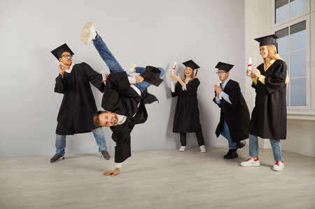 Congrats, grad. Group of excited university graduates in hats and robes dancing and having crazy fun on graduation day. Happy male student doing a break dance handstand and his friends are applaudingの写真素材