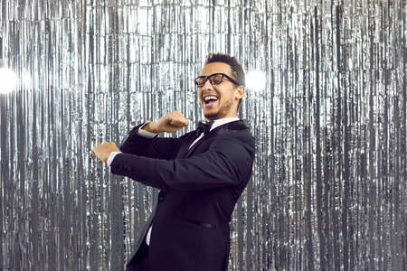 Young positive dark-skinned man in classic black suit having fun on silver background with shiny decorations. Disco banner with happy guy dancing and laughing out loud looking at camera. Party conceptの写真素材