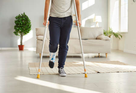 Closeup crop of unhealthy injured man with bandage on foot walk on crutches at home. Unwell guy have...