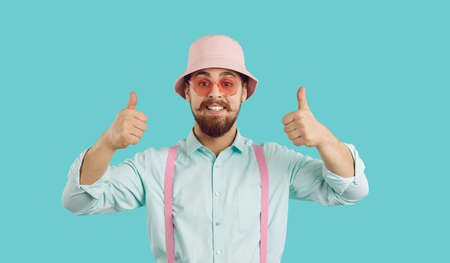 Cheerful funny charismatic young man showing thumbs up isolated on turquoise background. Bearded hipster in shirt, suspenders, panama and sunglasses looking at camera with funny expression on his faceの写真素材