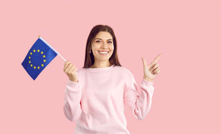 Symbol of Europe, EU association. Caucasian woman holding small flag with european union emblem isolated on pink background. Happy and smiling woman holding flag and pointing finger at copy space.の写真素材