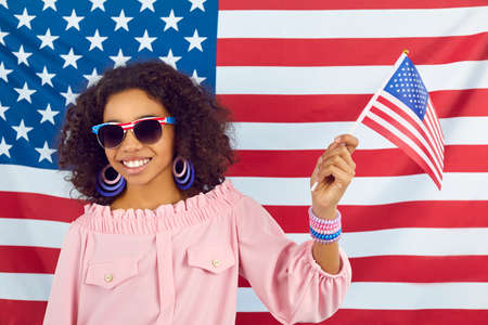 Afro American kid with flag of United States. Happy Black child celebrating Independence Day. Pretty girl in sunglasses standing against stars and stripes, holding little US flag and smilingの写真素材