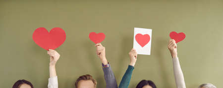 Unidentified men and women with raised hands hold models of hearts. Paper red hearts of different sizes in hands of unrecognizable people on khaki background. Concept of love, charity and volunteeringの写真素材