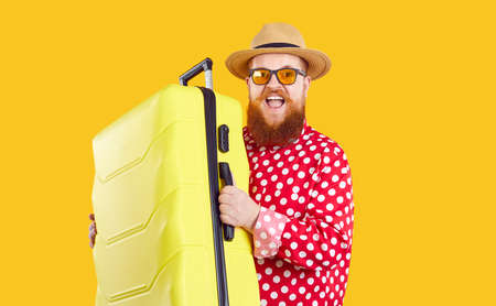 Joyfully funny excited extravagant bearded fat man with suitcase ready for summer vacation. Stylish chubby guy in red shirt, glasses and straw hat smiling holding travel suitcase on orange background.の写真素材