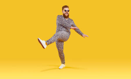 Plus size guy in funny pyjamas having fun in modern studio. Happy carefree fat man wearing comfortable leopard PJs and sunglasses dancing isolated on yellow background. Crazy party and fashion conceptの写真素材