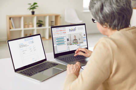Adult woman working on two computers. Entrepreneur sitting at desk in front of two laptop screens,...
