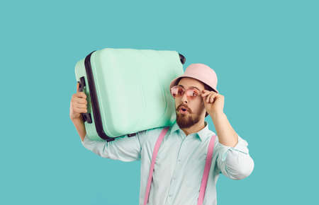 Amazed young man in summer hat and sunglasses isolated on blue studio background carry suitcase for vacation. Stunned guy ready for summertime trip or holidays. Travel and tourism concept.の写真素材