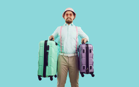 Embarrassed tourist guy holding two too heavy travel suitcases isolated on light blue background. Stylish man in panama and suspenders holds overweight luggage and looks at camera with funny grimace.の写真素材