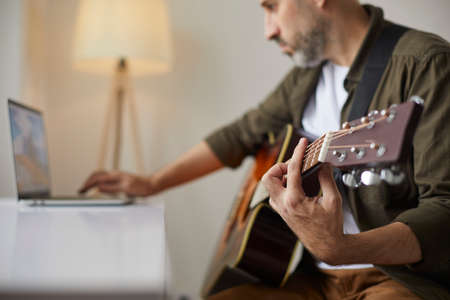 Man with acoustic guitar clamps desired chord while watching online lesson on laptop. Guitar fretboard and hand of man learning to play guitar using online tutorials. Selective and soft focus.の写真素材
