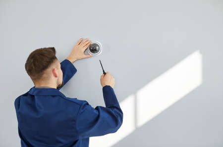 Male technician installing surveillance camera on light copy space wall. Back view of repair service worker using screwdriver to fit screws and adjust wall mounted CCTV security dome cam inside houseの写真素材