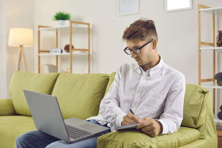 Serious young man using laptop for studying. College student in glasses sitting on sofa at home,...