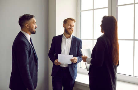 Business colleagues in formal suits have pleasant conversation during break in office. Two men and woman working in same company discuss documents and business projects standing by bright window.の写真素材