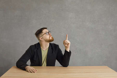Smart young Caucasian man shows a thumbs up as a sign that he has a great innovative idea. Guy sits at the table on a gray background and looks up. Gray background. Place for text.の写真素材
