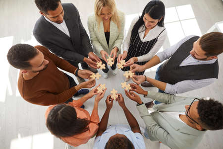 Business people hold puzzle pieces as sign of team building, company merger or joint venture. Top view of men and women standing in circle and connecting puzzles. Concept of teamwork and partnership.の写真素材