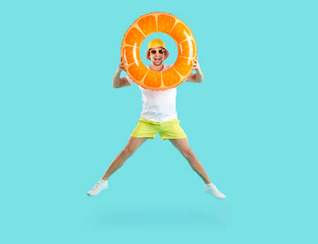 Summer. Cheerful funny man looking through inflatable circle bouncing with him light blue background. Joyful playful guy in shorts, T-shirt and panama has fun with swimming circle with orange designの写真素材