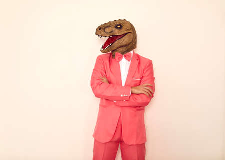Studio shot of strange guy wearing wacky ugly dinosaur mask. Man in pink party suit, bow tie and funny silly scary masquerade lizard mask standing with his arms folded isolated on white backgroundの写真素材