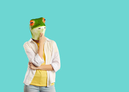 Funny woman in rubber mask of frog stands in pensive pose on turquoise background. Casual woman with frog head stroking chin thinking and looking at copy space. Advertising creative banner.の写真素材