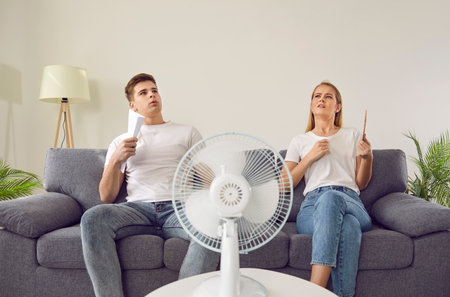 Young couple at home suffering from hot sultry sweltering summertime weather. Sweaty exhausted people waving sheets of paper sitting on sofa with electric fan in overheated house. Summer heat conceptの写真素材