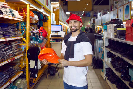 Shopping in hardware store. Man during shopping chooses protective hard hat in hypermarket of building materials. Portrait of smiling satisfied male shopper holding hard hat standing in aisle of storeの写真素材