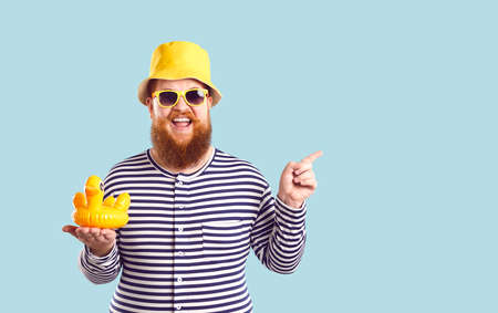 Funny fat man with inflatable duck in his hand points to copy space on pastel light blue background....