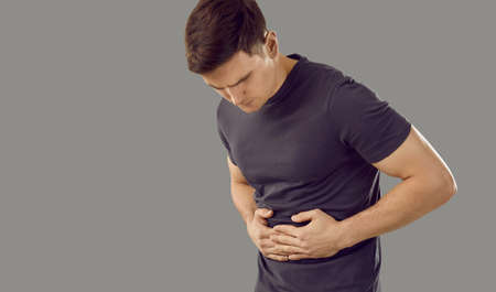 Sick young man isolated on grey studio background struggle with stomachache. Pain or spasm in abdomen. Unwell ill guy suffer from ache in belly. Gastrointestinal problem and indigestion concept.の写真素材
