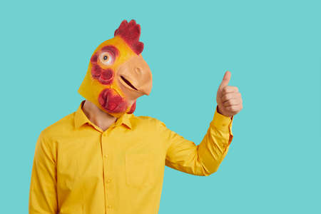 Funny man with chicken head raises his thumb up demonstrates you OK gesture, consent or his support. Eccentric man in yellow shirt and with rubber mask of chicken on his head on light blue background.の写真素材
