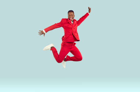 Happy funny young african american man in red suit high jumps on pastel light gray background. Joyful crazy man in red suit and white sneakers jumps happily in air. Banner. Full length.の写真素材