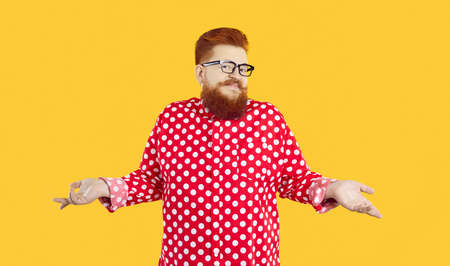Funny stylish bearded fat man who knows nothing or doubts and therefore shrugs isolated on orange background. Red-haired chubby man spread his arms to side shows that he has no idea what is going on.の写真素材