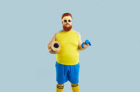 Funny redhead bearded overweight man in sportswear and glasses isolated on blue studio background hold sport equipment ready for workout. Fat male lose weight motivated to train and exercise.の写真素材