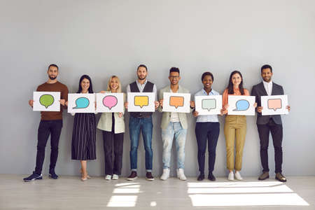Everyone has their own opinion. Portrait of group of men and women holding white sheets of paper with empty colored speech bubbles on them. Multiracial people standing near wall and empty room.の写真素材