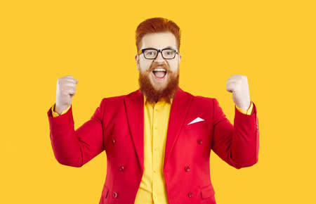 Funny excited fat guy in red suit and glasses celebrating great success. Happy overjoyed euphoric businessman with ginger beard celebrating, having fun, raising up fists and shouting Yesの写真素材