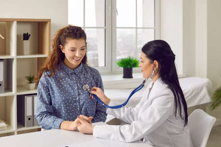 Young woman comes to clinic for heart and lungs checkup. Friendly female doctor sitting at desk in...
