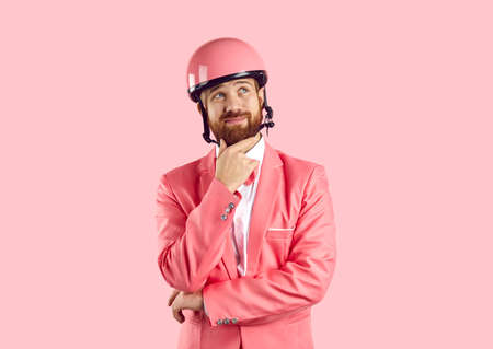 Funny young Caucasian man in pink helmet and suit isolated on studio background think or ponder. Cool male in costume and headpiece consider offer or sale deal. Advertising concept.の写真素材