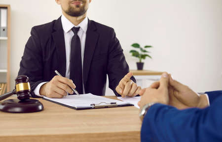 Lawyer meeting client in his office. Cropped shot of law specialist in suit sitting at desk with documents, holding pen, and giving consultation to young man. Legal advice, lawyer services conceptの写真素材