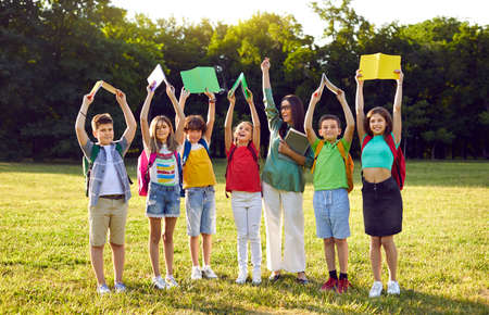 Female teacher and students with books having fun interesting class outside classroom. Portrait of happy, excited school teacher together with children standing in row in green field or schoolyardの写真素材