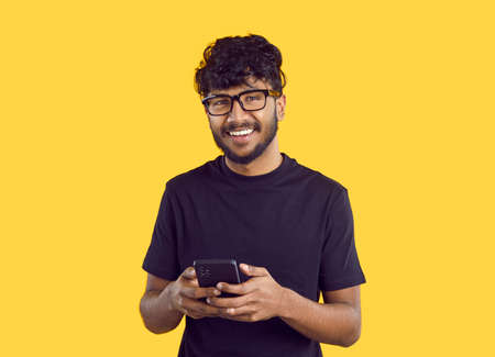 Crop shot of smiling millennial Indian guy in glasses isolated on yellow studio background using modern smartphone. Happy young biracial man text or message online on cellphone. Communication.の写真素材