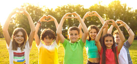 Children show gesture of love. Group of joyful preteen kids standing in sunny park with arms raised show heart shape made by hands. Boys and girls in colorful summer clothes express love. Web banner.の写真素材