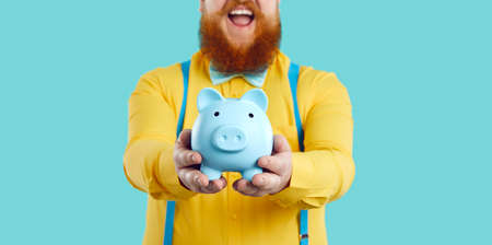 Piggy bank in form of blue pig in hands of joyful contented unrecognizable bearded fat man. Man in yellow shirt and suspenders holding piggy bank on light blue background. Close up. Cropped image.の写真素材