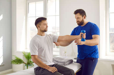 Professional physiotherapist controls his patient who is doing arm muscle rehabilitation exercises. Serious young man doing physical exercises with dumbbells at modern physiotherapy clinic or centerの写真素材